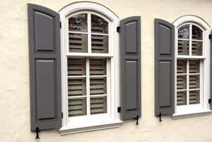 PROPERLY INSTALLED GRAY ARCH TOP PANEL SHUTTERS ON TAN STUCCO HOME